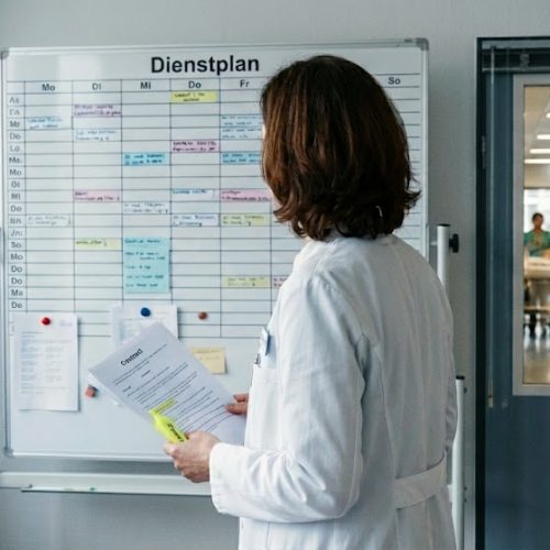 Person in weißem Arztkittel betrachtet einen großen Dienstplan an einer Tafel, im Hintergrund durch eine Glastür sichtbarer Klinikflur mit Pflegepersonal und Patientinnen.